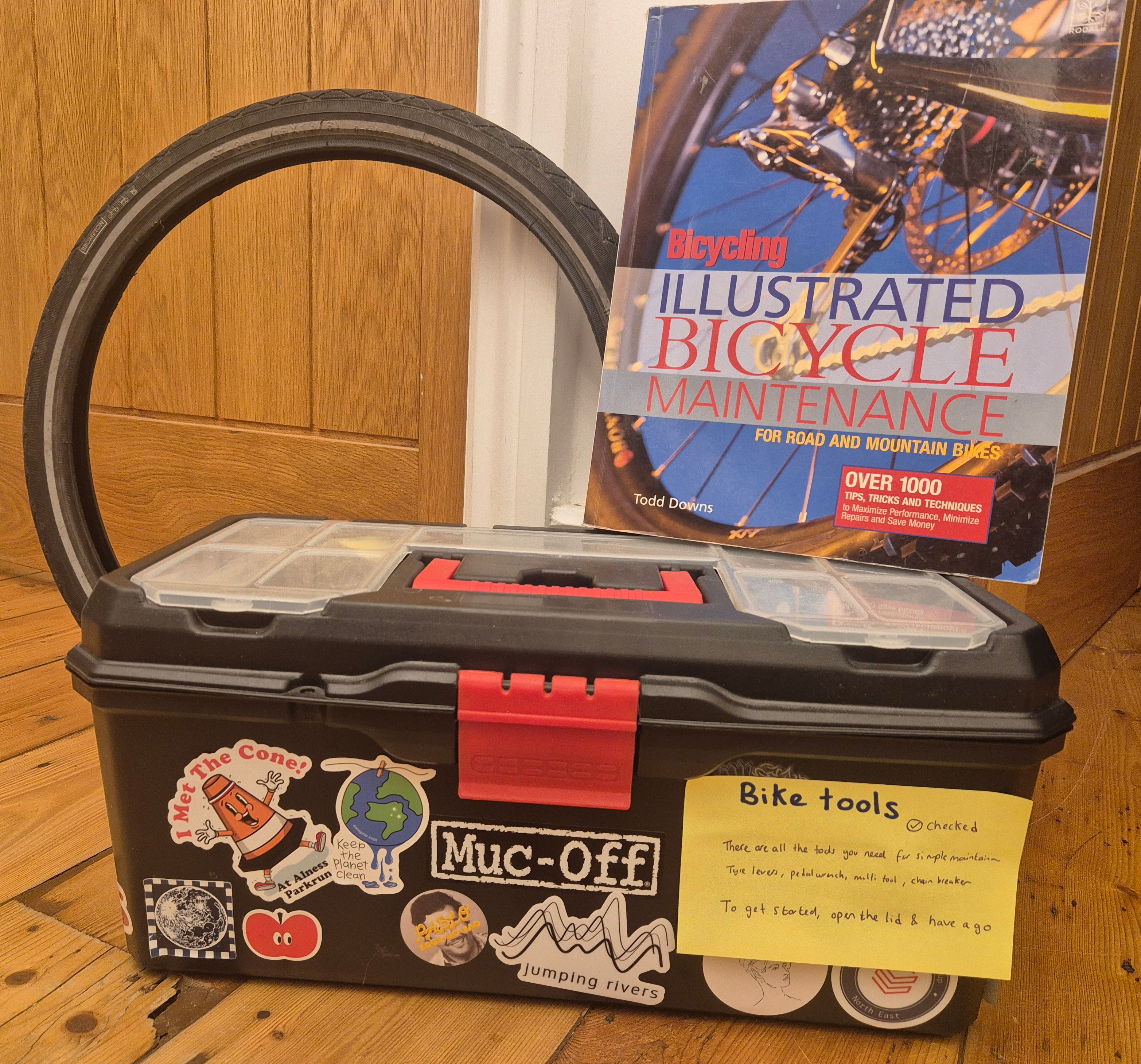 The closed toolbox has a label on the side stating the toolbox has been checked and providing a longer description of what to use the toolbox for. Behind the toolbox is a spare tyre and an illustrated book of bicycle maintenance.
